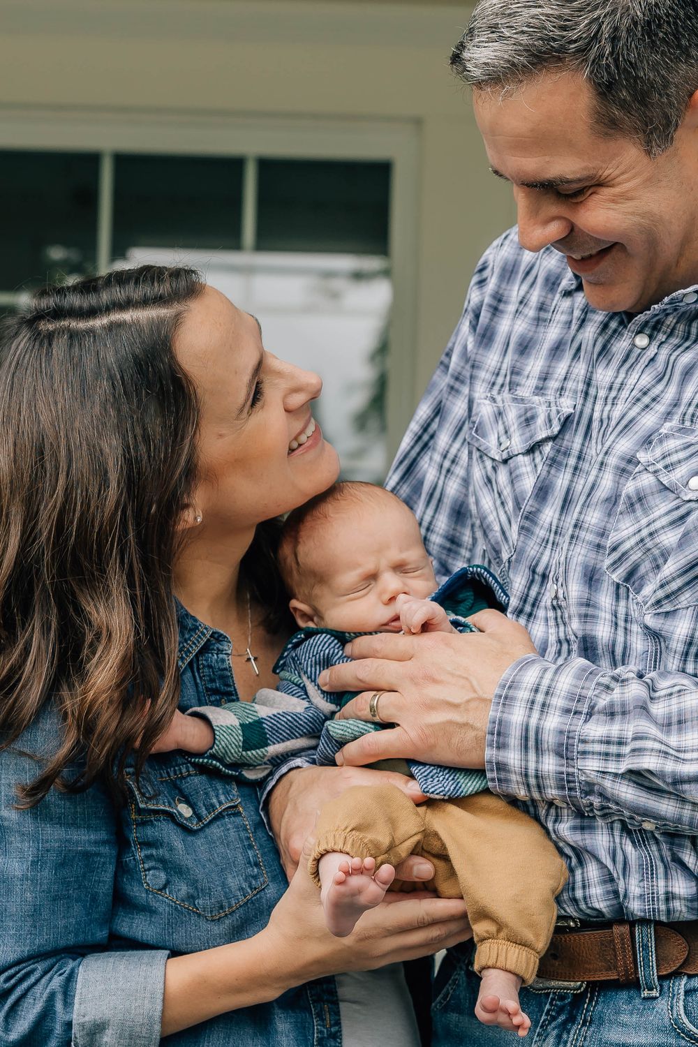 Happy couple smiles lovingly at their sleeping newborn baby held in fathers arms on a porch.