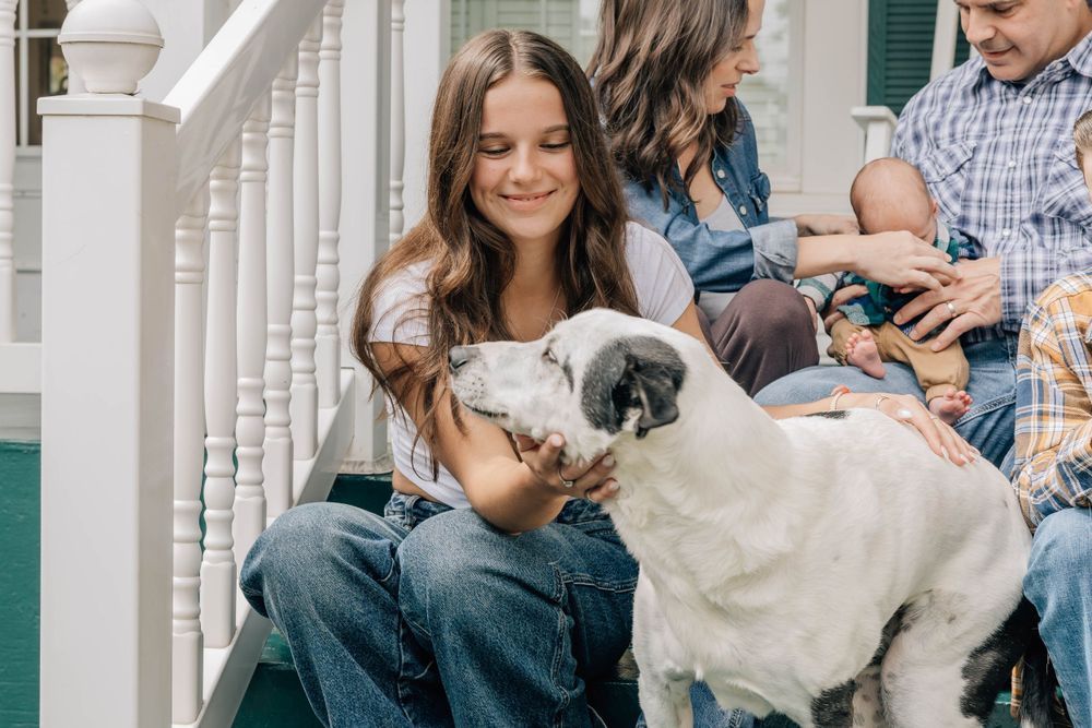 Young girl smiles while petting a large white dog on porch steps with family in background.