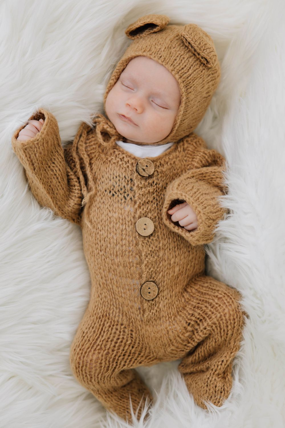 Sleeping newborn baby dressed in a brown knit bear onesie and matching hat lying on a white fluffy rug.