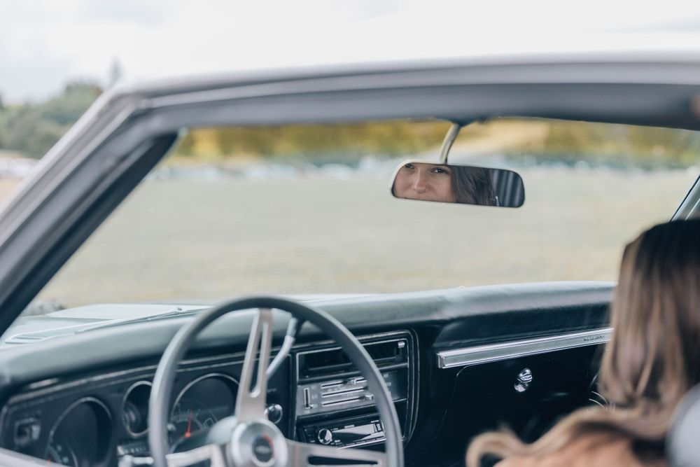 Interior view of a classic vintage car dashboard with a rearview mirror reflection while a woman sits in the passenger seat.