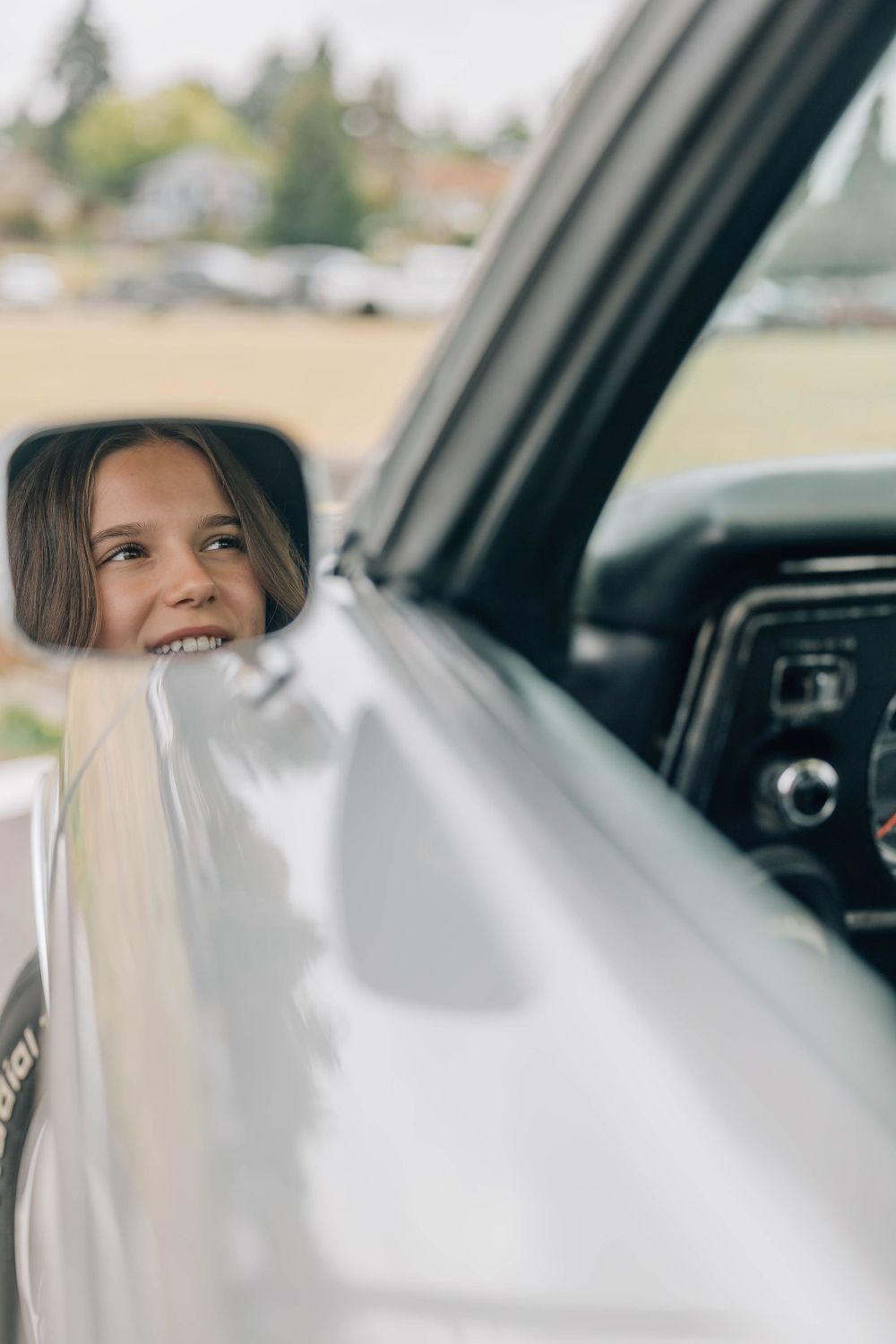 Smiling young girl with long brown hair reflected in the side mirror of a modern silver truck outdoors.