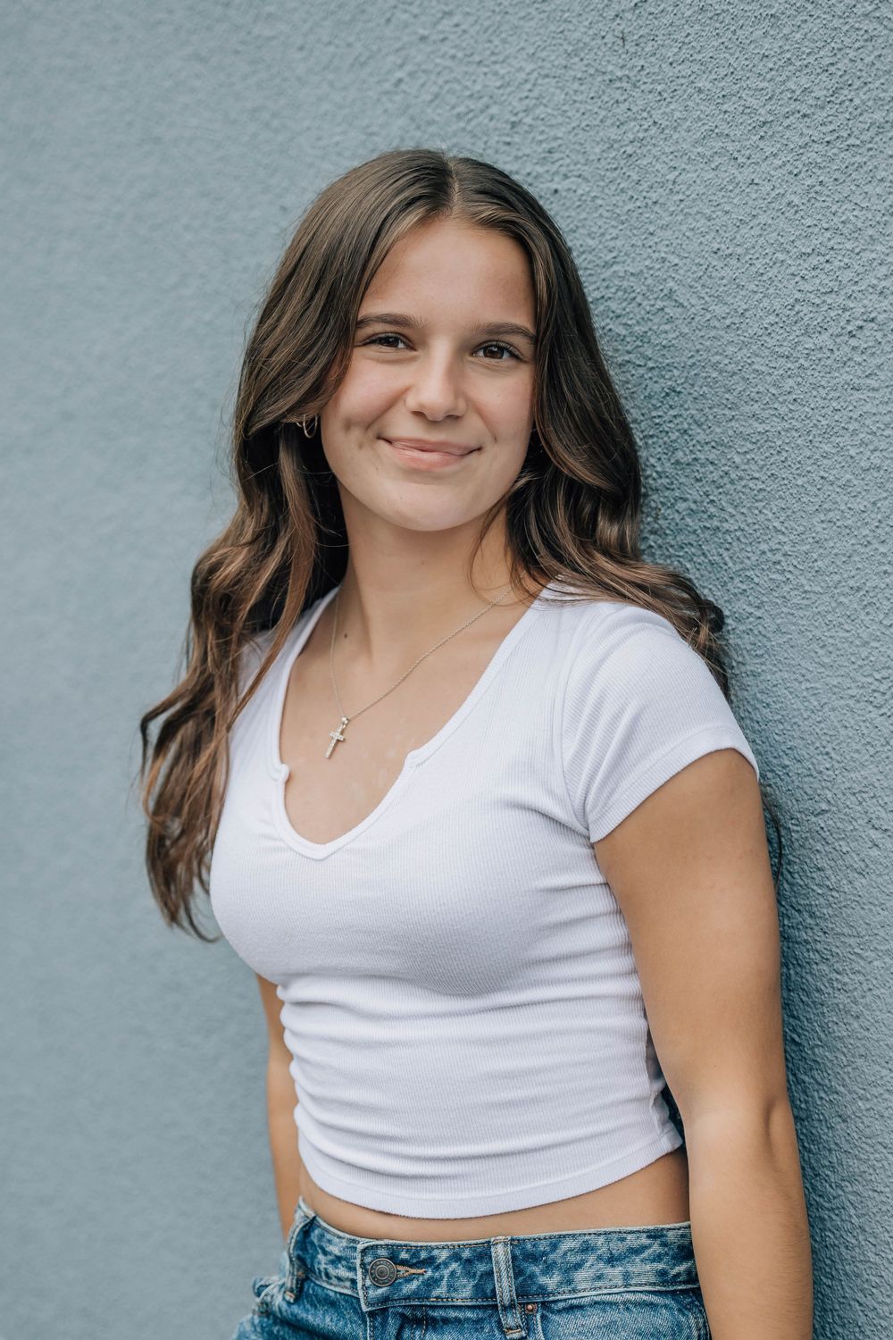 Smiling teenage girl with long wavy brown hair wearing a white crop top and jeans leaning against a gray wall.