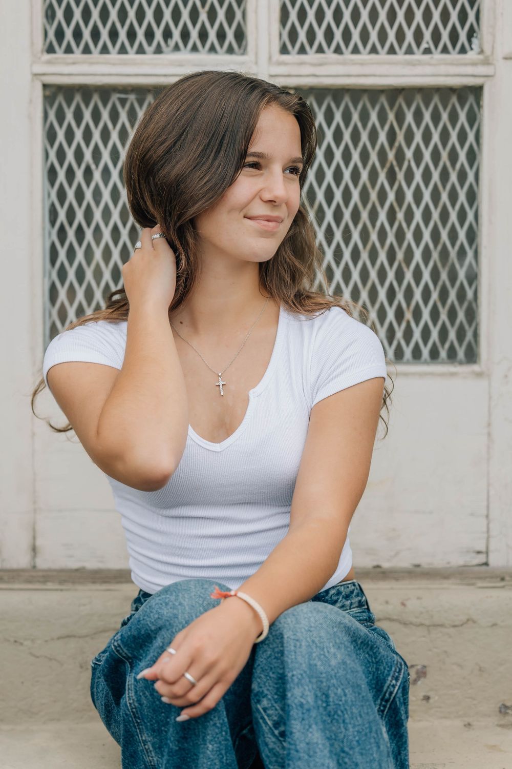 Young woman in white t-shirt and jeans sitting outdoors, touching her hair and smiling near a lattice window.