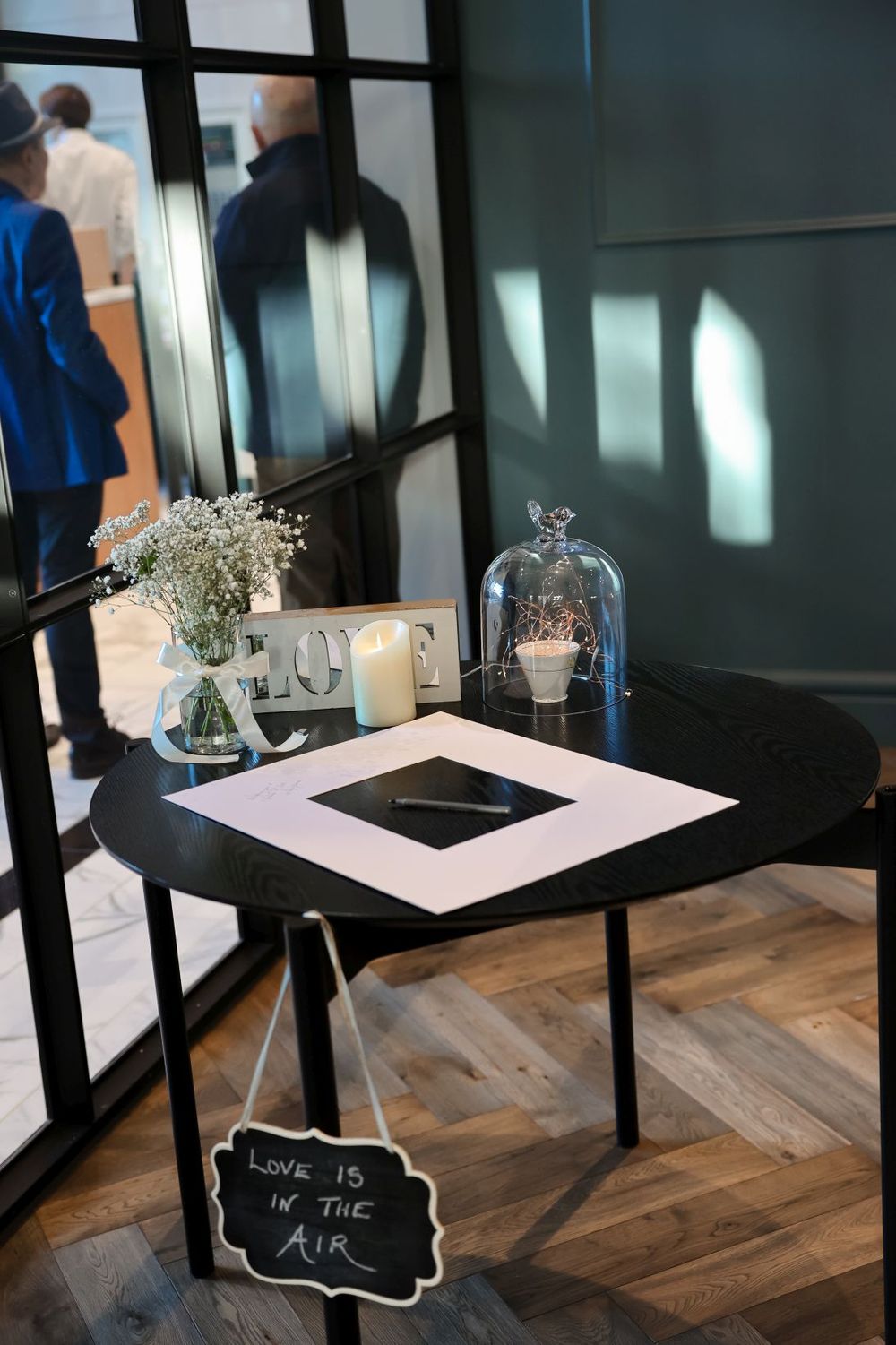 A modern wedding venue table setup with a black frame, glass cloche, and baby's breath centerpiece on hardwood floors.