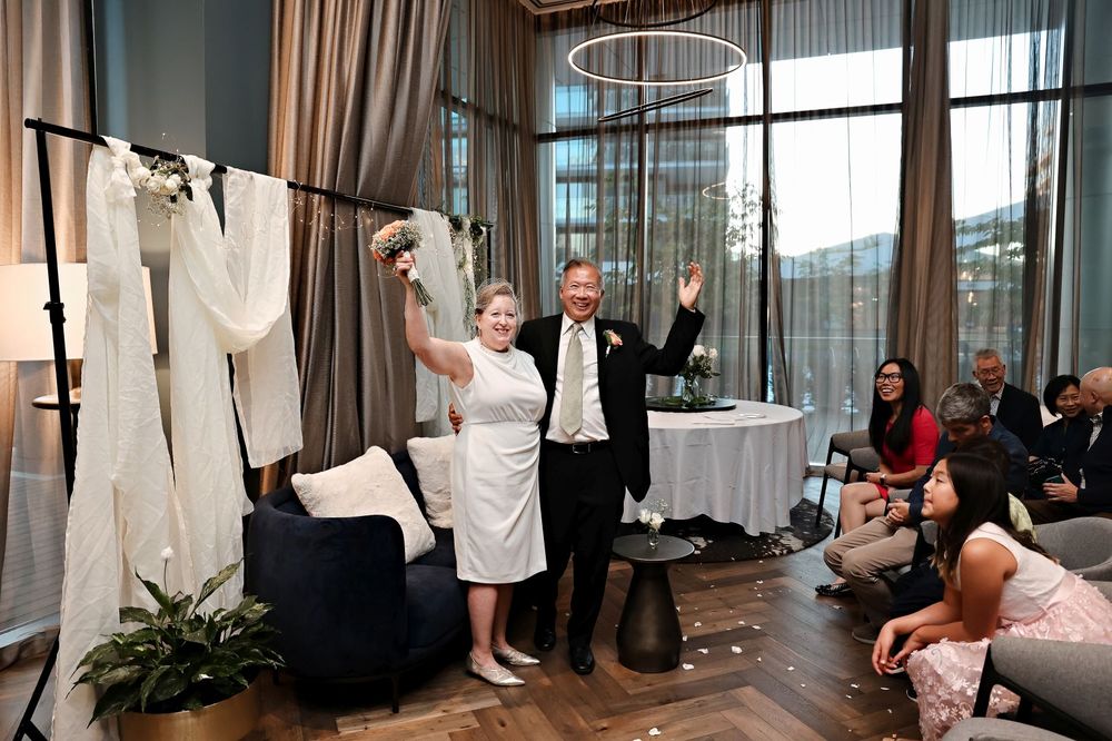 A couple celebrates joyfully at their wedding reception in a modern venue with floor-to-ceiling windows.