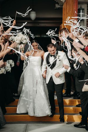 Newlyweds exit church amid ribbon streamers, groom in white tuxedo, bride in halter wedding gown.