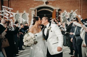 Bride and groom kissing outside brick church with guests waving white ribbon streamers in celebration.