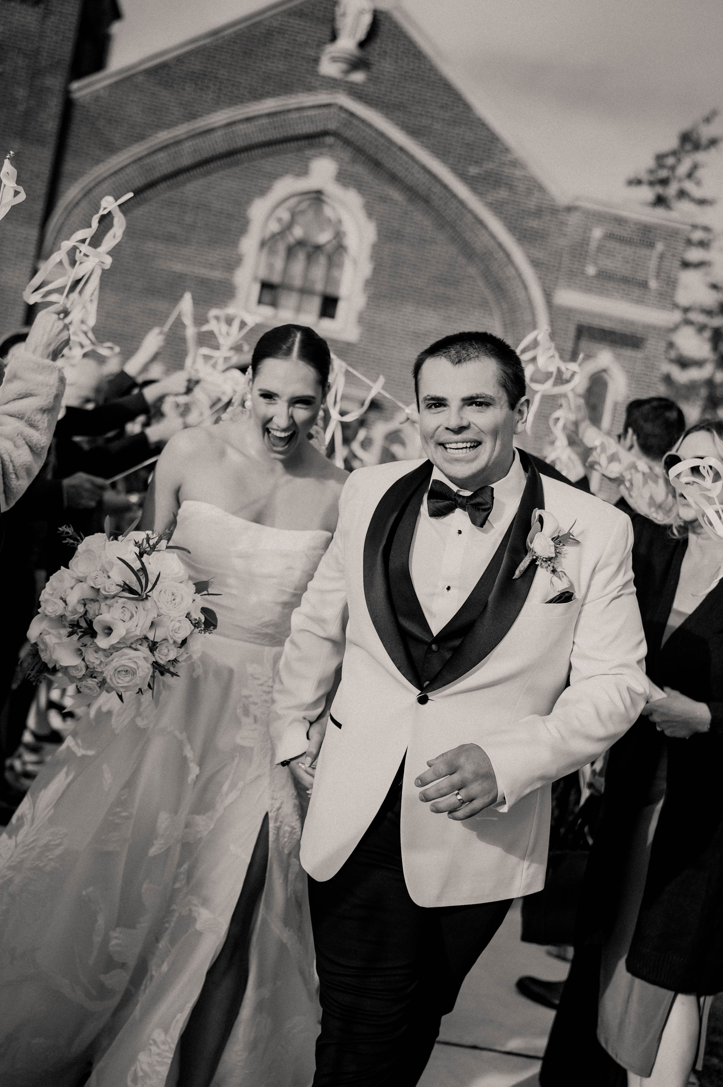 Black and white photo of smiling newlyweds exiting church with guests waving streamers in celebration.