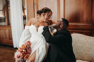 A bride in a white off-shoulder gown laughs with her groom seated on a vintage sofa indoors.