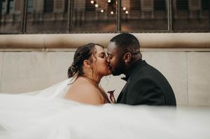 A bride and groom share a passionate kiss beneath a flowing veil outside a stone building.