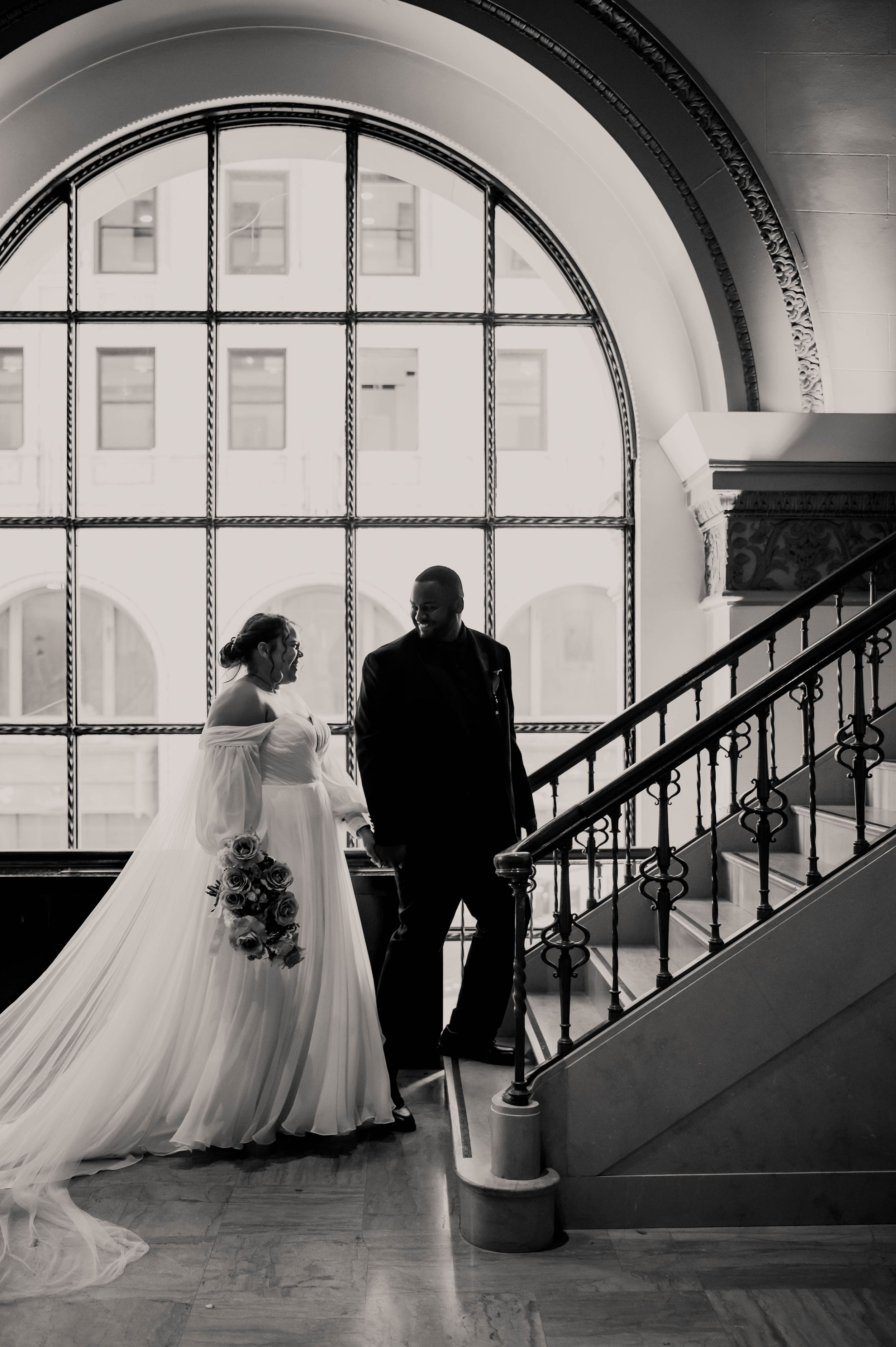 A black and white silhouette of a bride and groom standing by a grand arched window on a staircase.