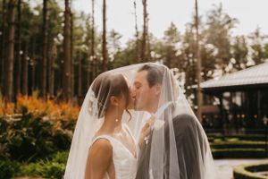 Newlyweds kiss romantically under a draped bridal veil surrounded by tall pine trees.