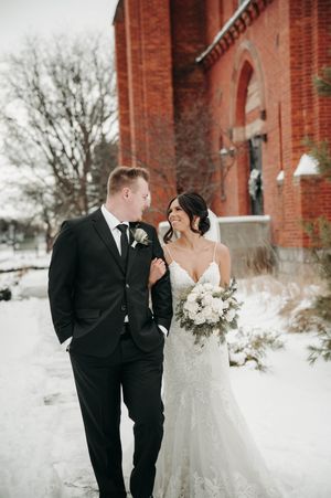 Bride in lace gown and groom in black suit walk together beside Abbey on Main in winter snow.