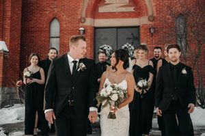 Wedding party in black attire walks together outside red brick church in winter snow setting.