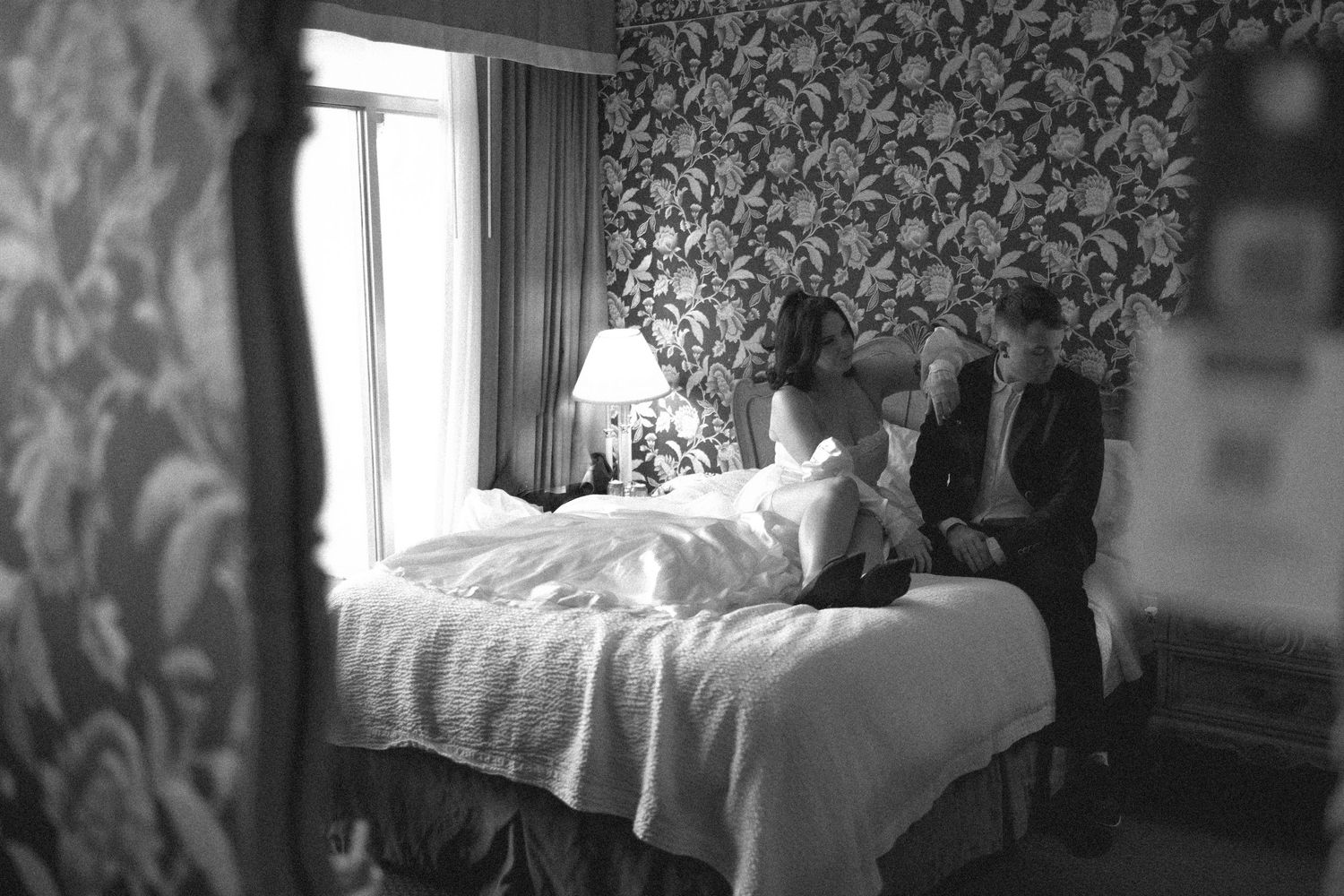 A couple sits together on a bed in a room with vintage floral wallpaper, captured in black and white.