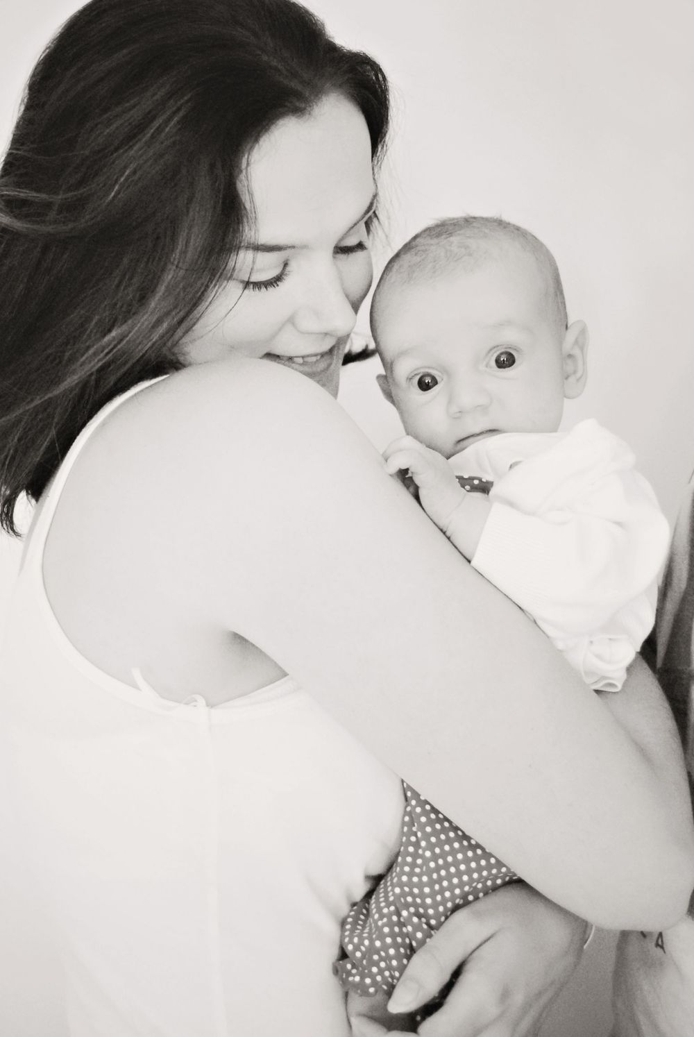 A tender black and white portrait of a mother embracing her baby in a white outfit.