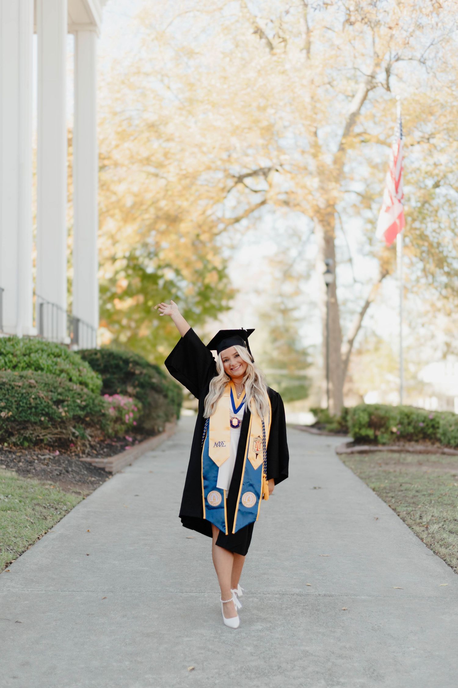 Graduate in cap and gown celebrating on campus walkway with autumn trees in background.