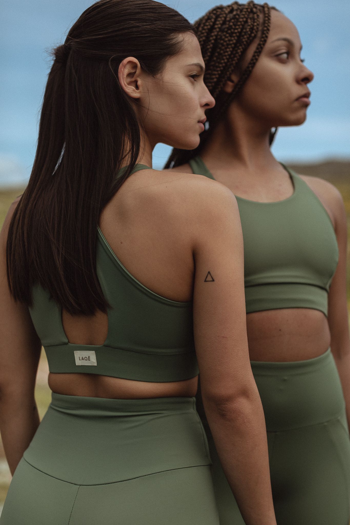 Two women in olive green athletic wear standing back to back in natural outdoor setting.