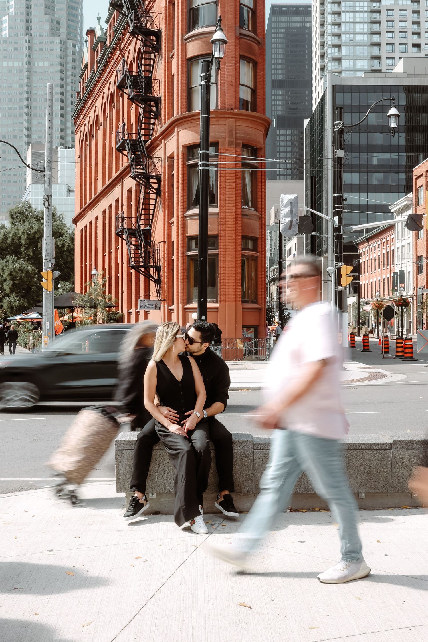 Couple sitting by historic red brick building as pedestrians walk past in urban setting.