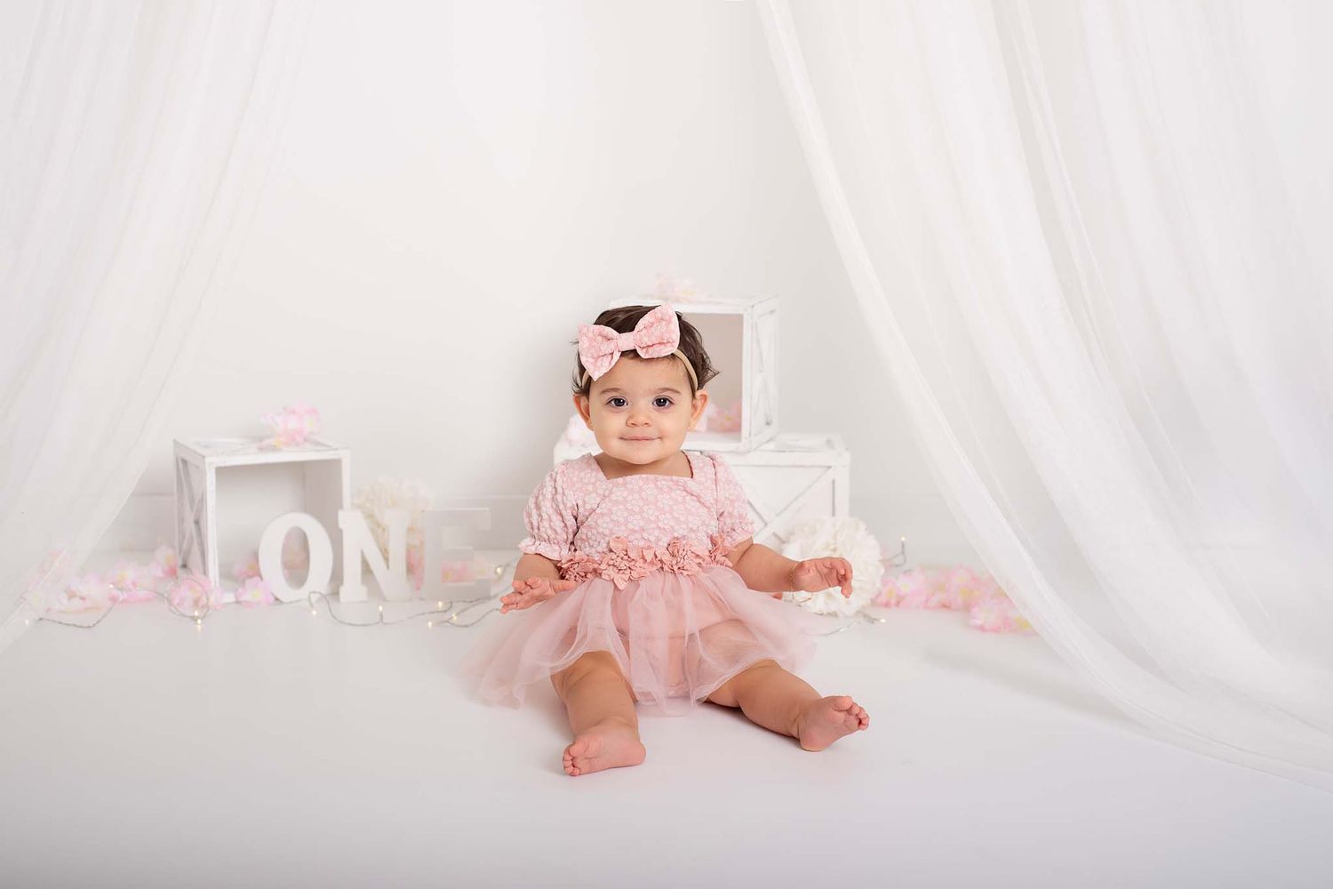 bright and white, toddler girl with pink dress smiling for her first birthday, with sheer curtains, pink flowers and white box decor