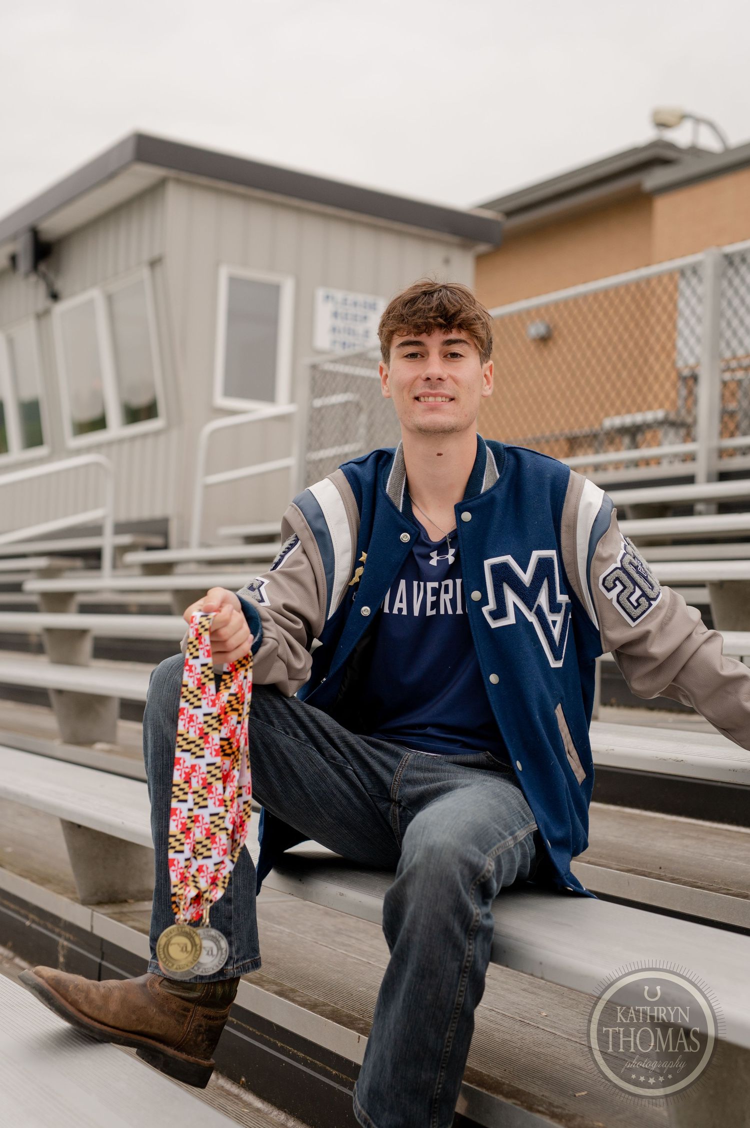 High school student with skateboard poses on bleachers wearing varsity jacket and jeans.