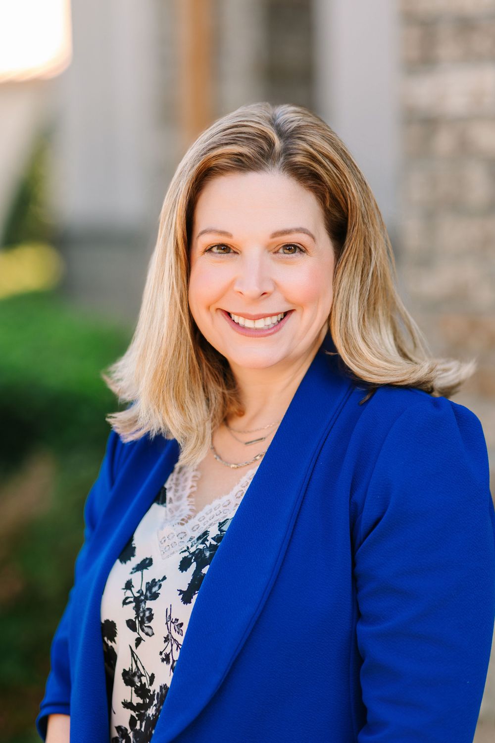 Professional headshot of a person in royal blue blazer and floral top outdoors.