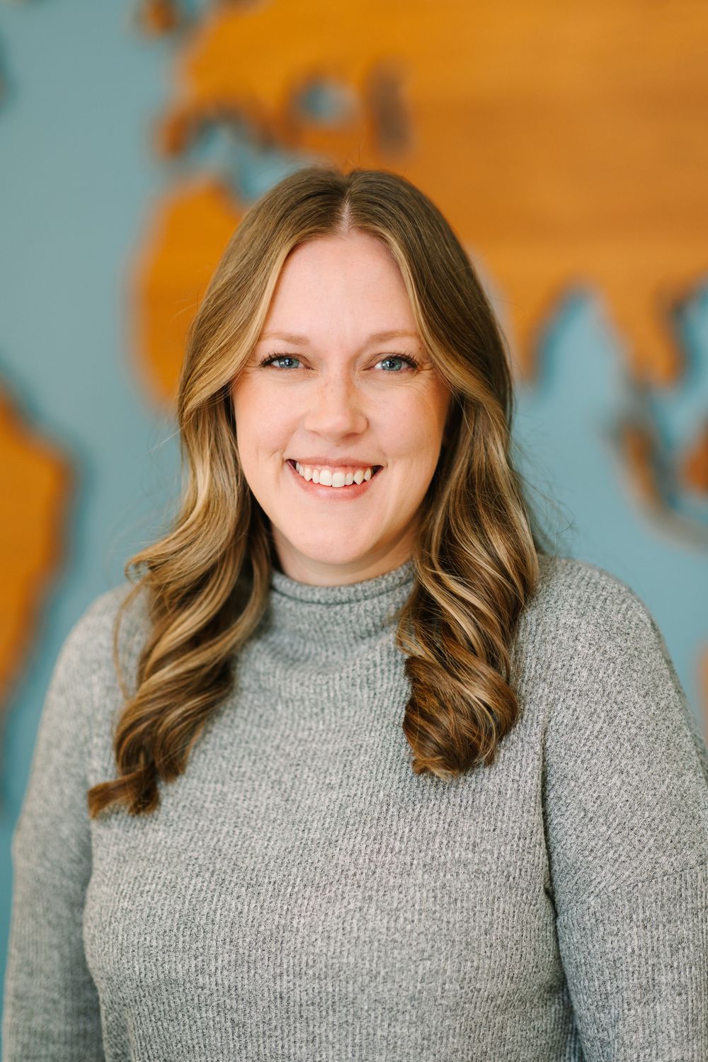 Professional headshot of a person in grey sweater with wavy hair against world map background.