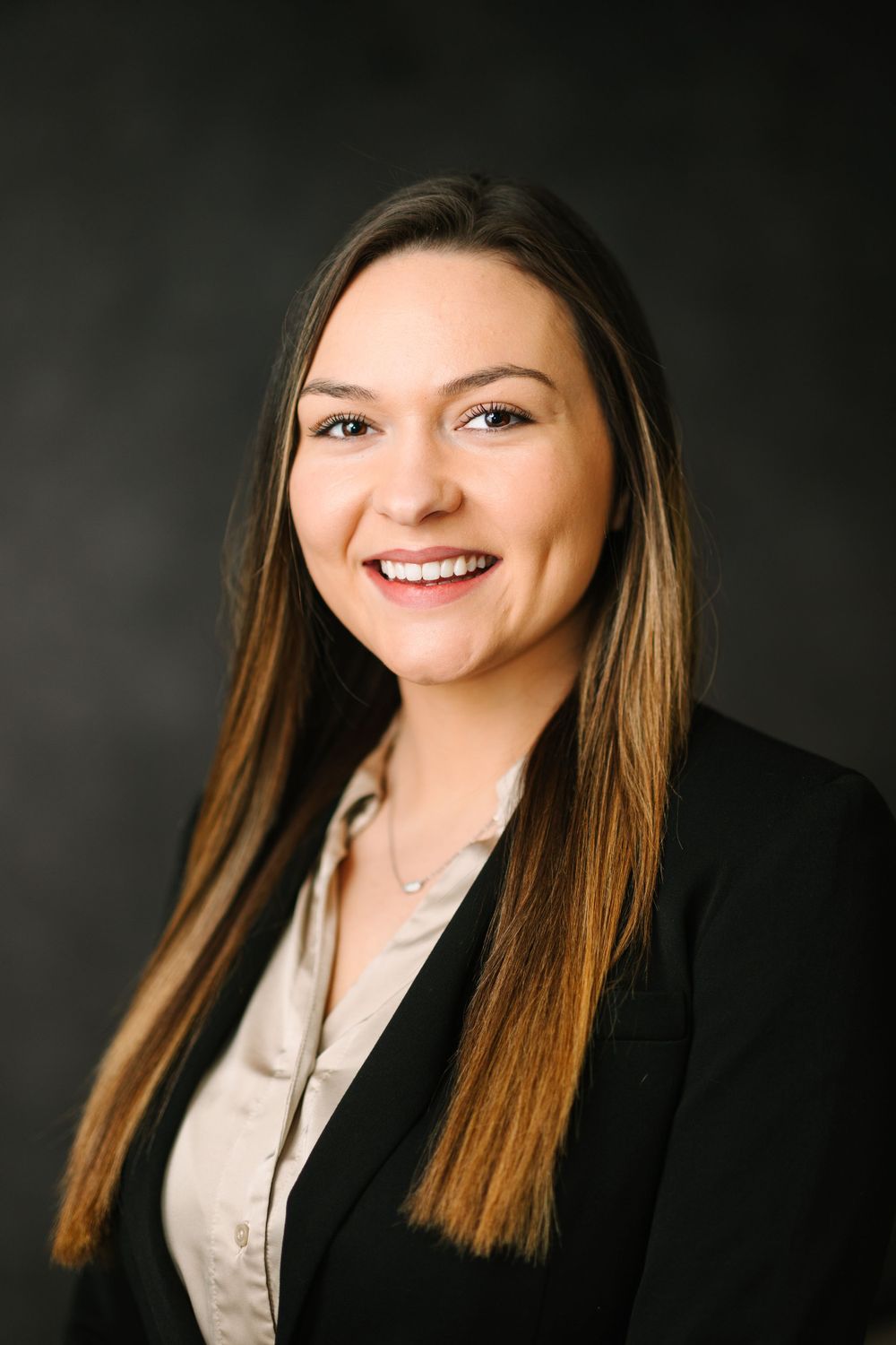 Professional headshot of a person wearing a black blazer and neutral top against dark background.