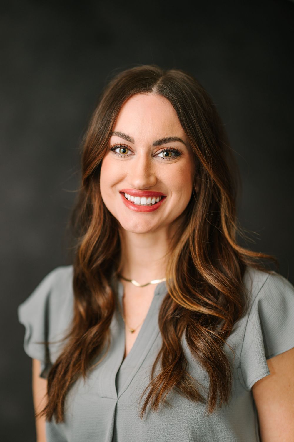 Professional portrait of a person wearing a grey blouse with wavy brown hair against dark backdrop.