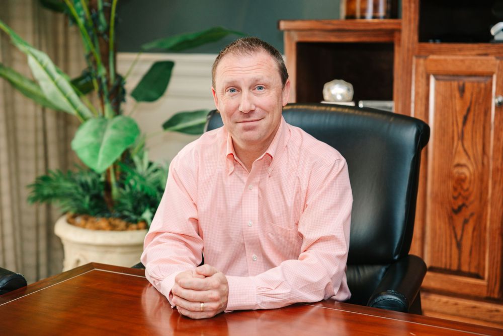 Professional portrait of a person in pink button-up shirt seated at desk with indoor plants.