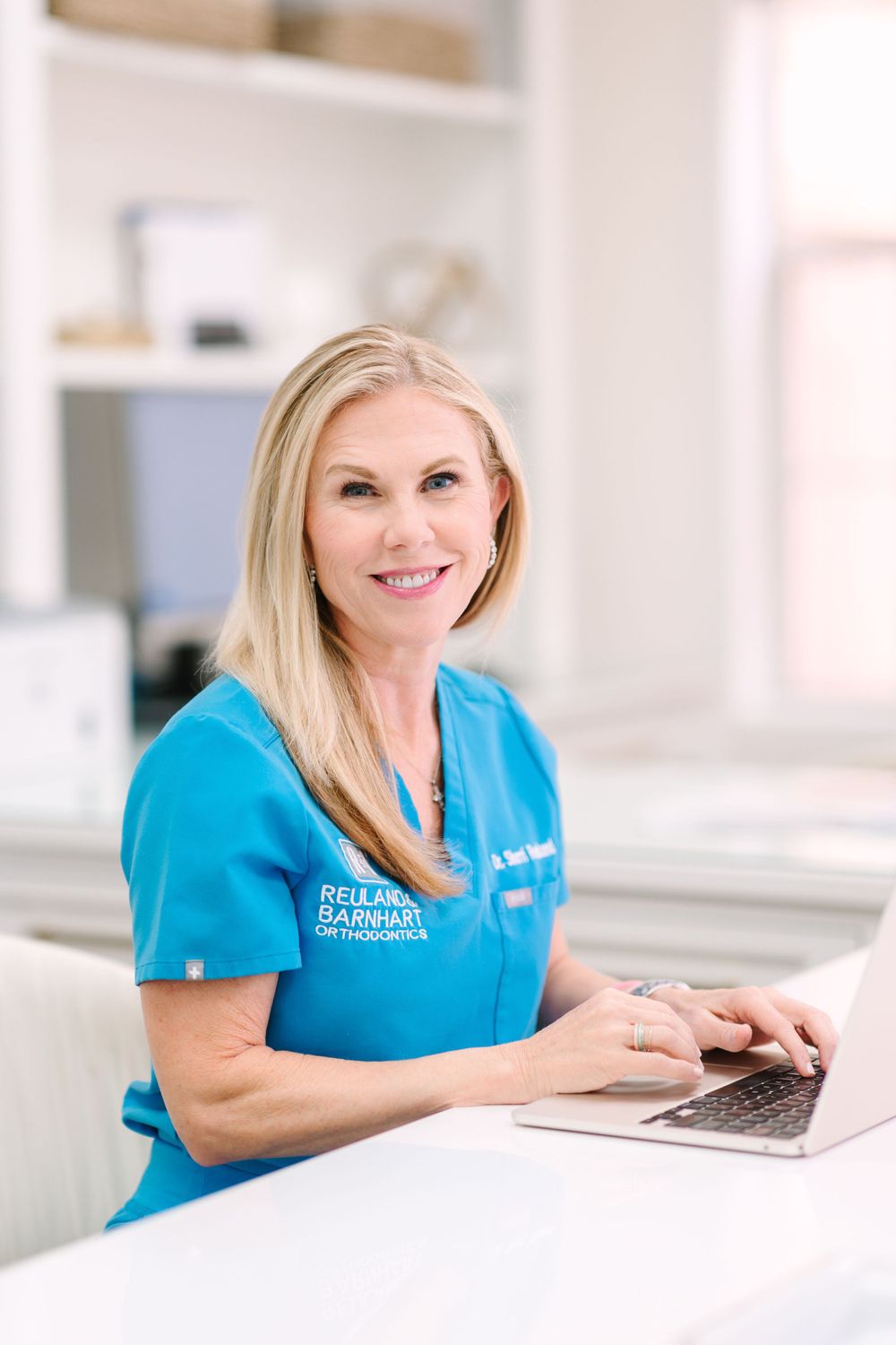 Medical professional in blue scrubs working at desk with laptop in bright office setting.