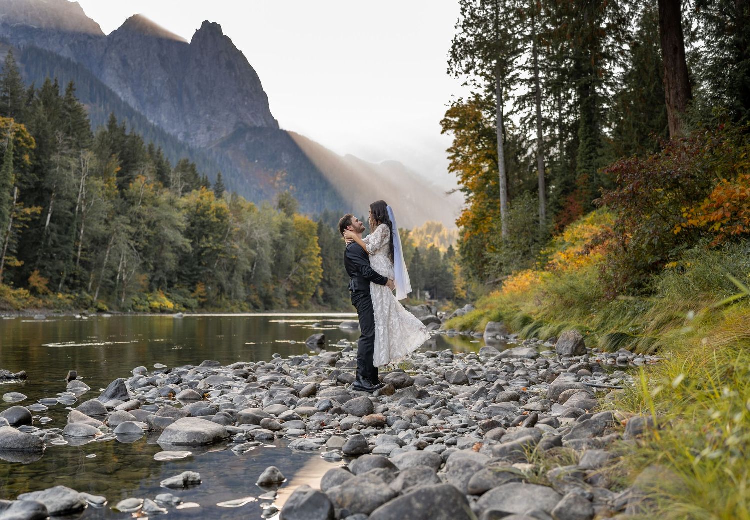 Couple in wedding attire embracing by a mountain river with dramatic landscape.