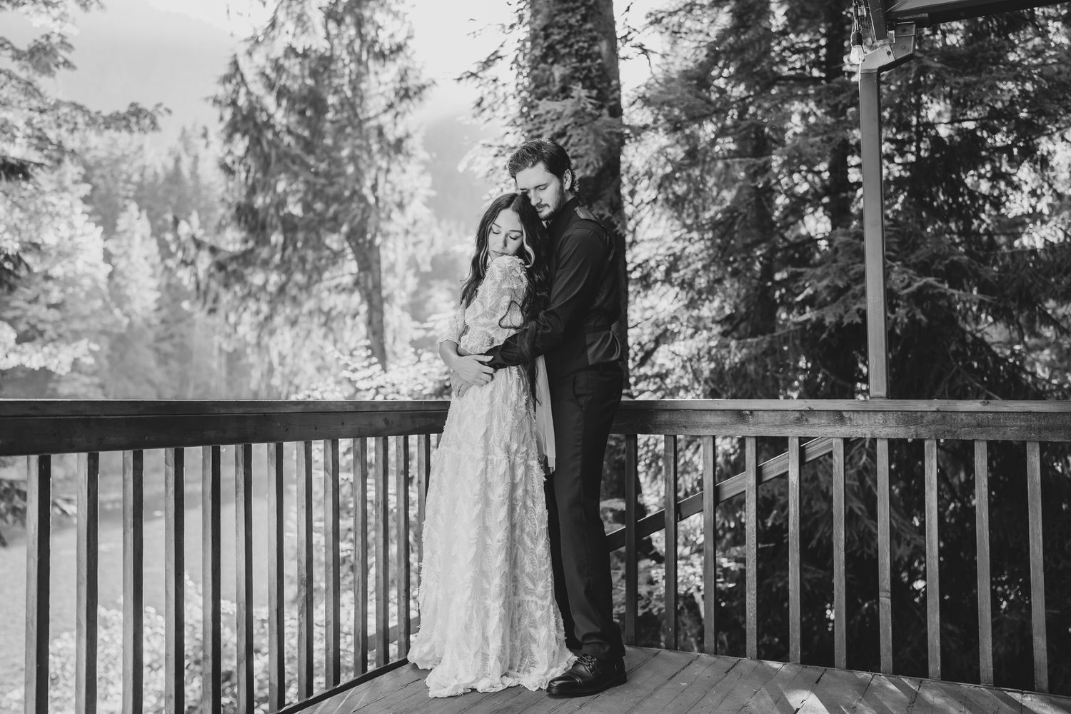 Black and white portrait of a couple on a wooden deck surrounded by evergreen trees.