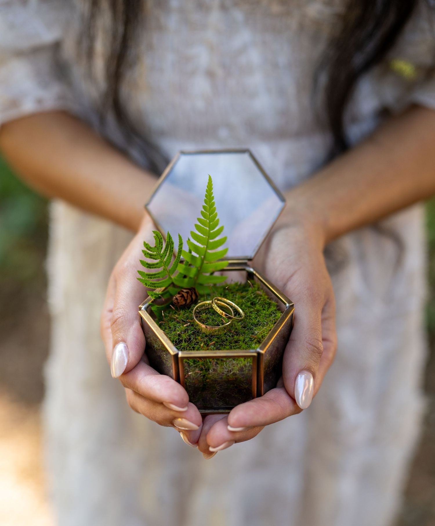 Hands holding a hexagonal glass box with ferns and wedding rings on moss.