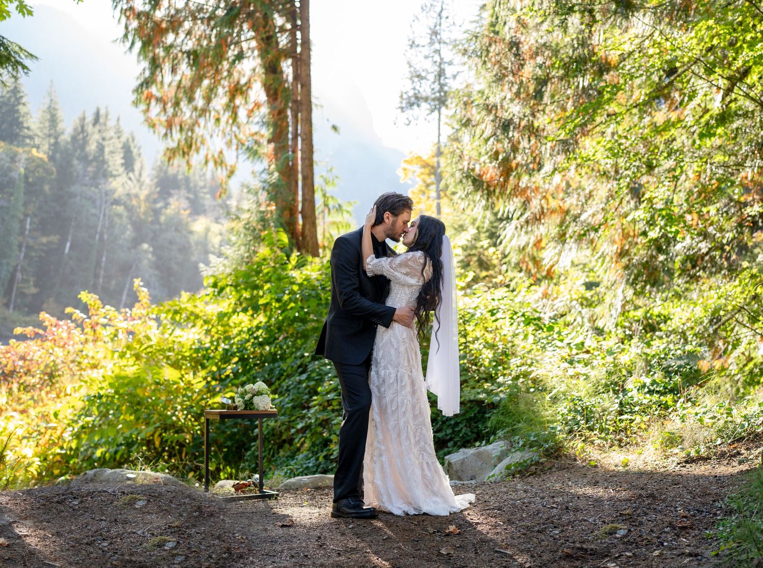 Newlyweds embrace in a romantic forest setting with sunlight filtering through trees.