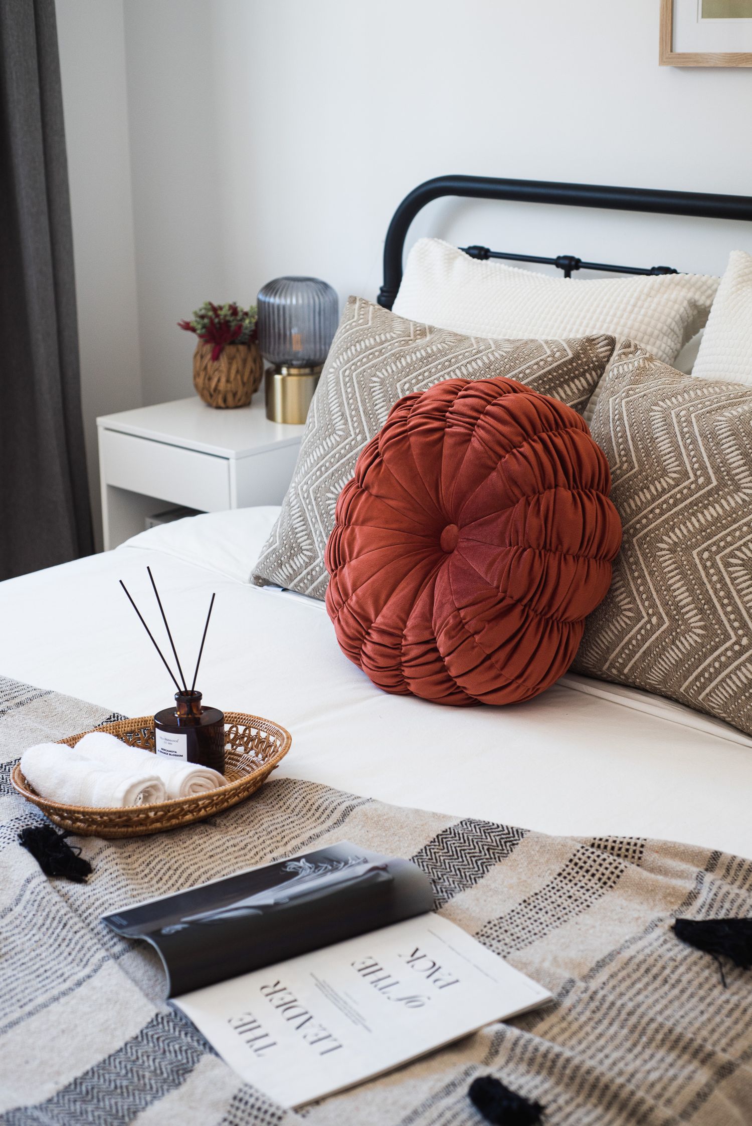 Cozy bedroom decor with rust round pillow, textured throw pillows and diffuser on white bedding.