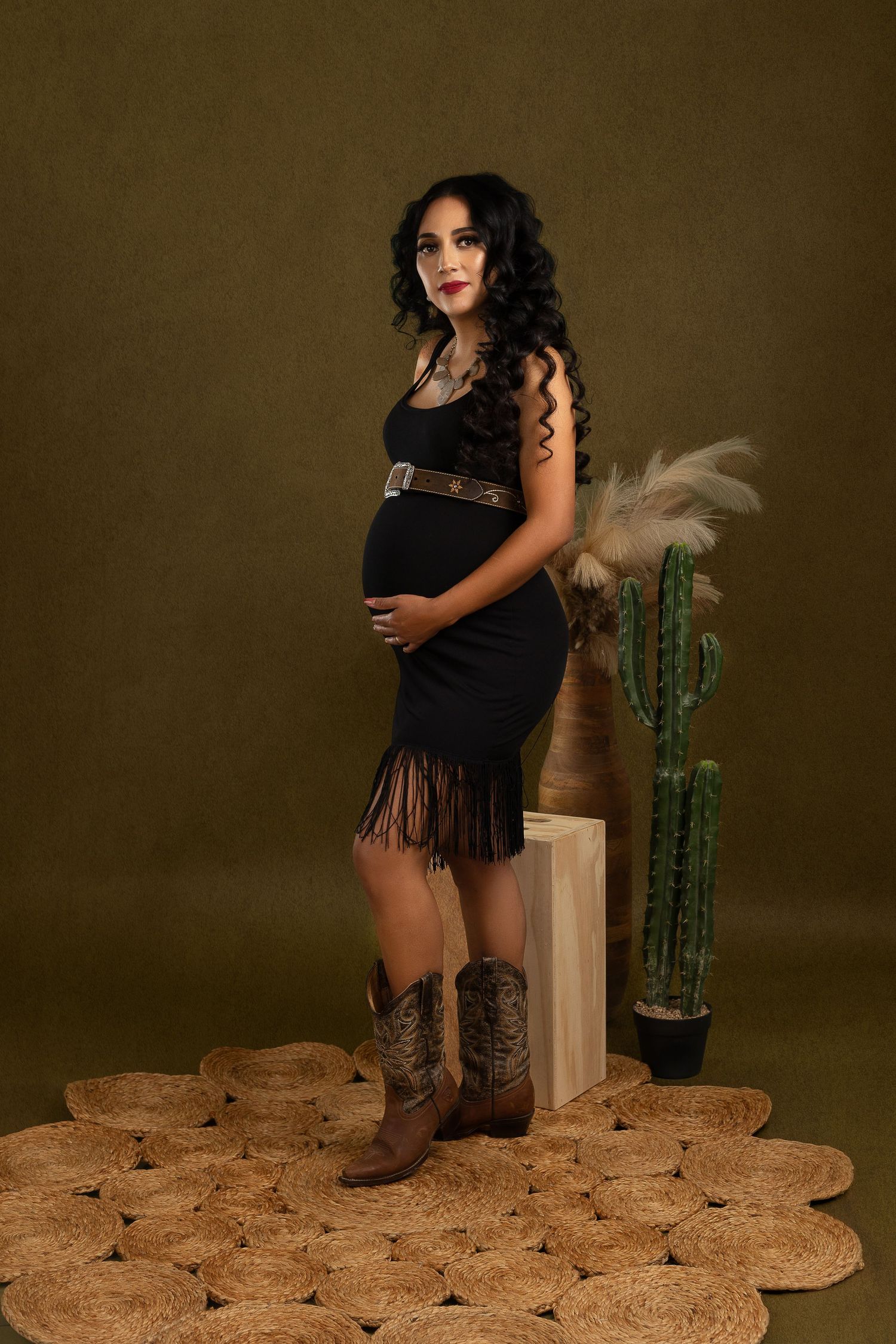 A maternity photo with Western style featuring a black fringe dress, cowboy boots, and desert-themed decor with cactus.