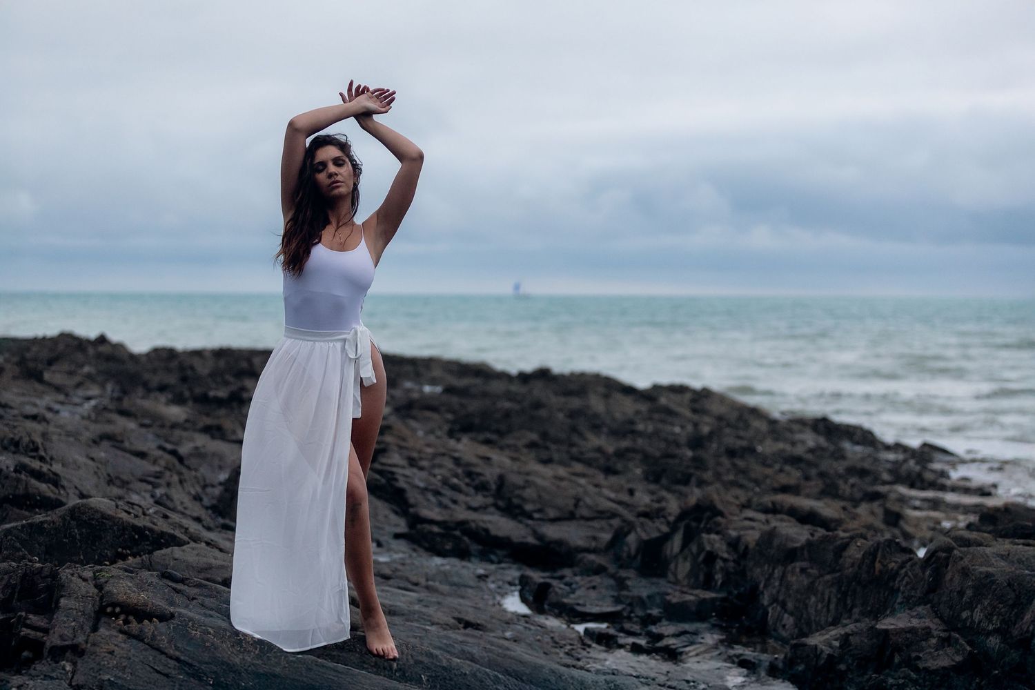 Artistic beach portrait of a dancer in black dress with flowing fabric against overcast sky.