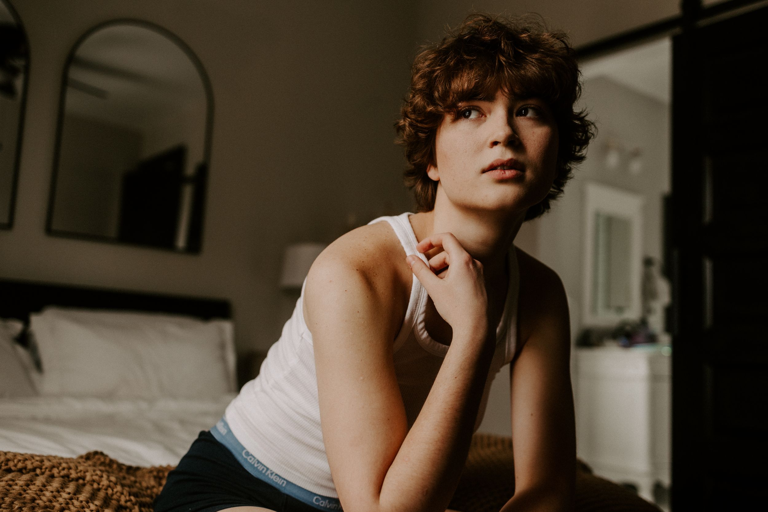 Moody indoor portrait of someone in white tank top on bed with arched mirror in background.