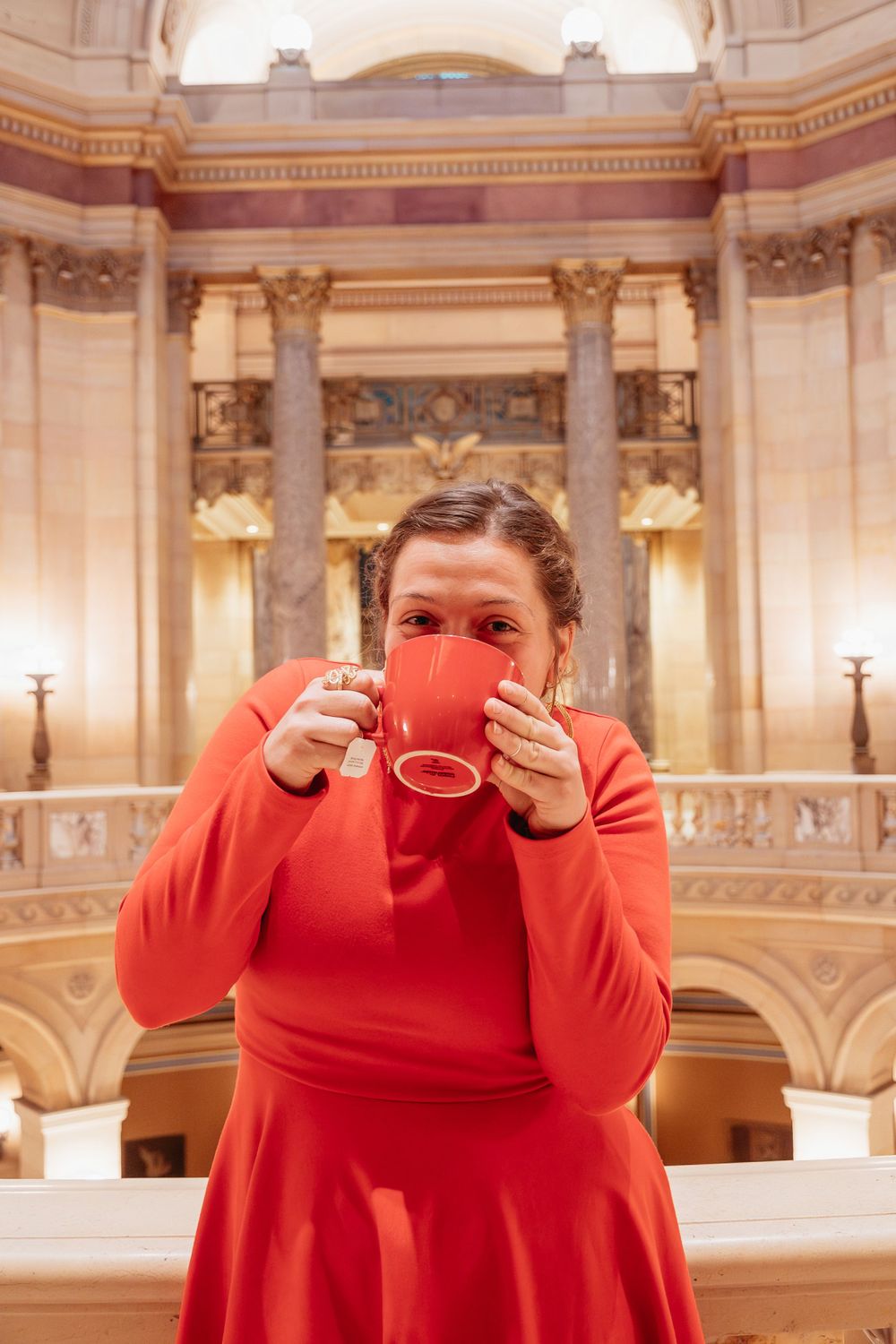 Person in a bright red dress sips from a cup inside an ornate building with classical columns and architecture.