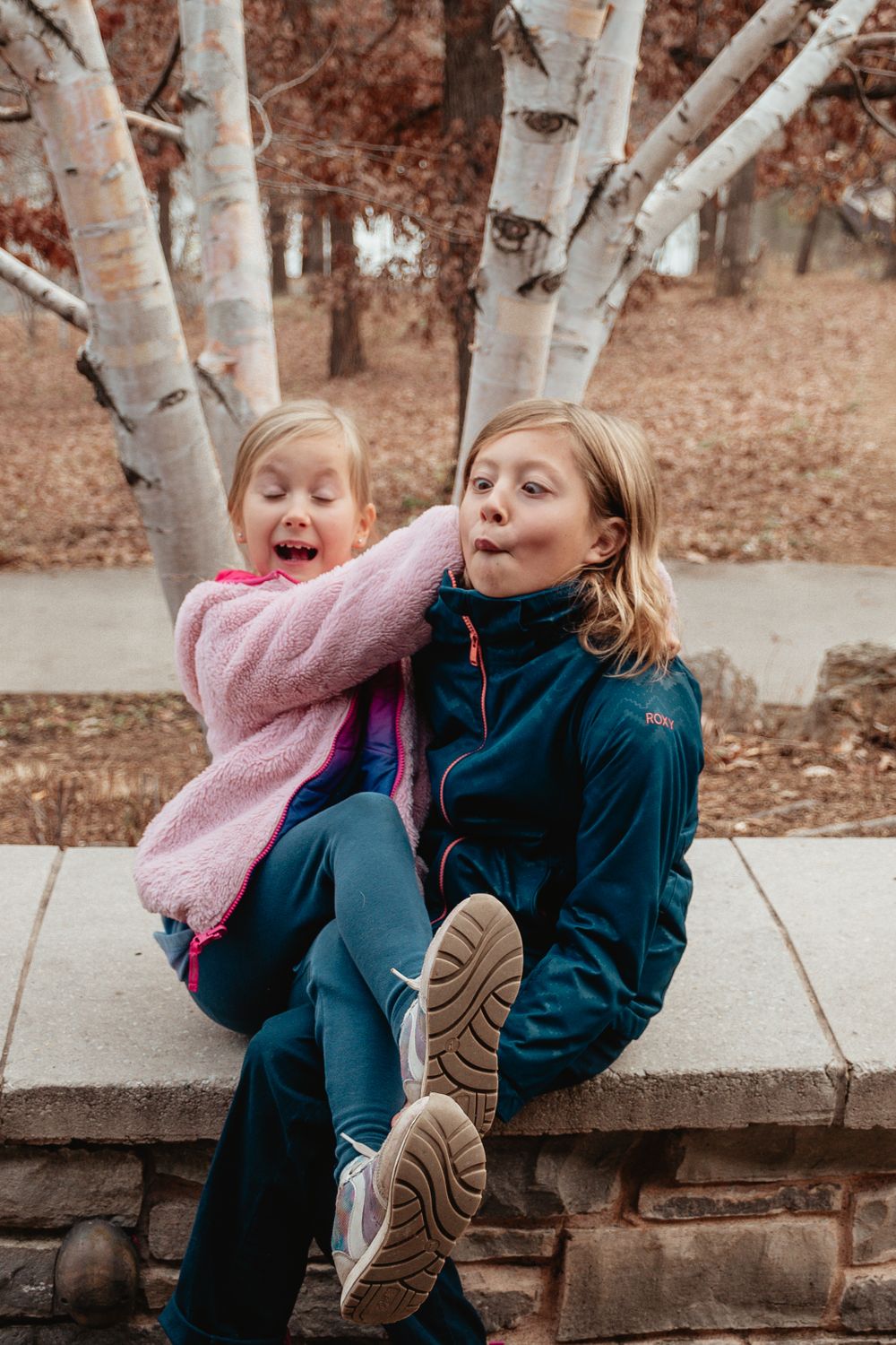 Two people sharing playful moment together on stone wall with birch trees in background.