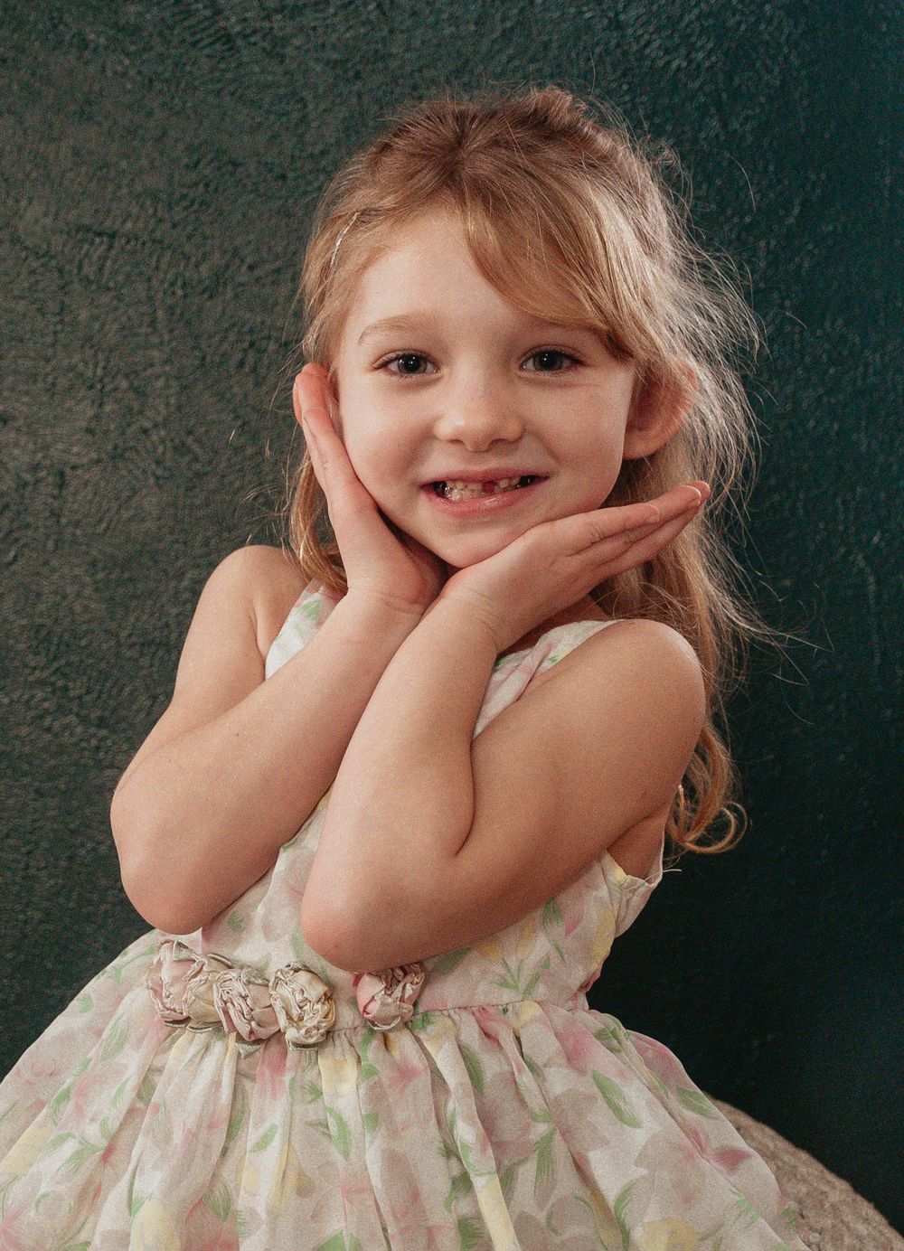 A child in a floral dress with a cream-colored sash poses with hands framing their smiling face against a dark green backdrop.