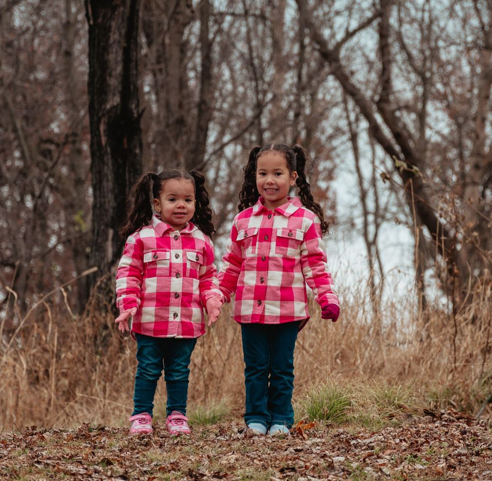 Two children wearing matching pink plaid jackets stand together on a wooded trail.