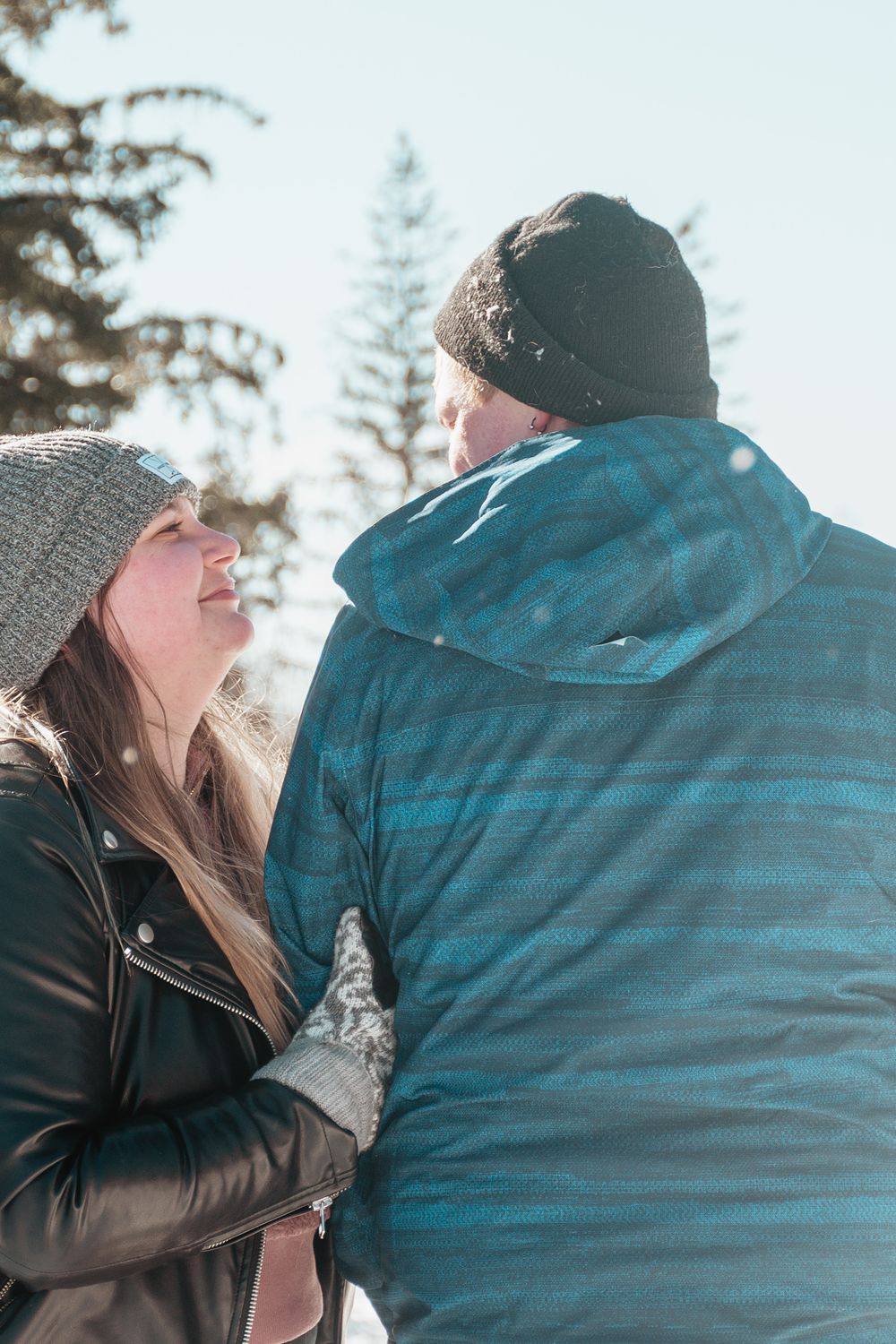 A couple in winter clothing share a candid outdoor moment against a backdrop of evergreen trees in soft sunlight.