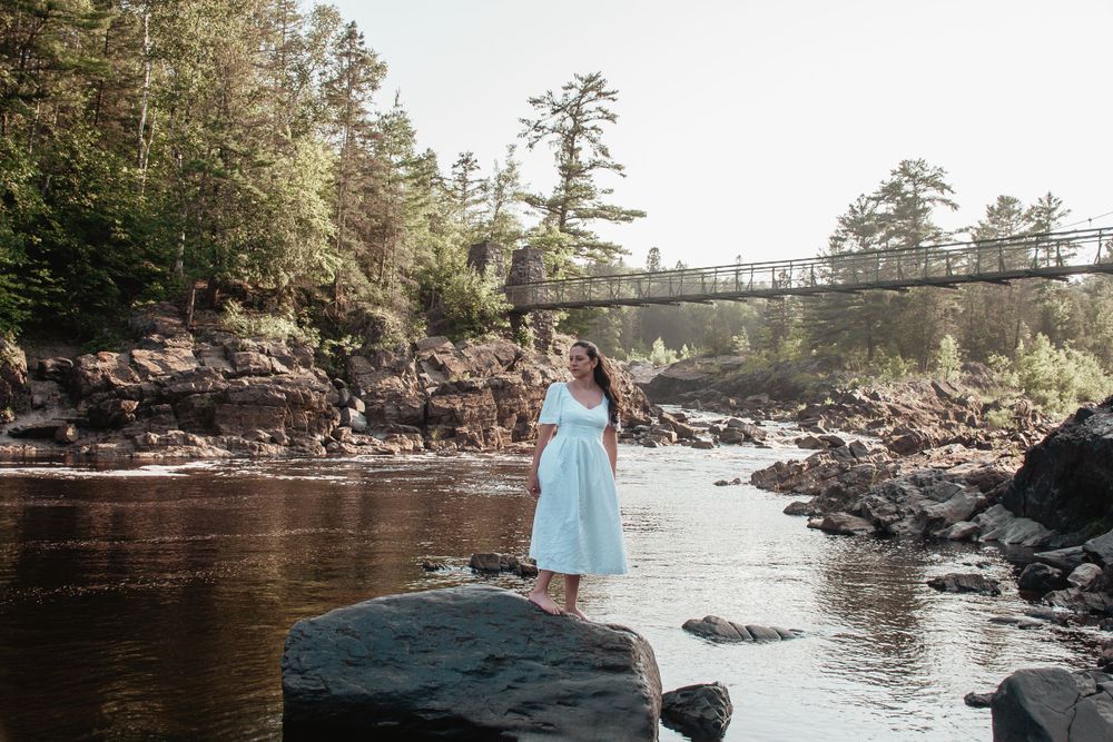A blue dress standing on rocks beside a rushing river with a bridge in the distance.