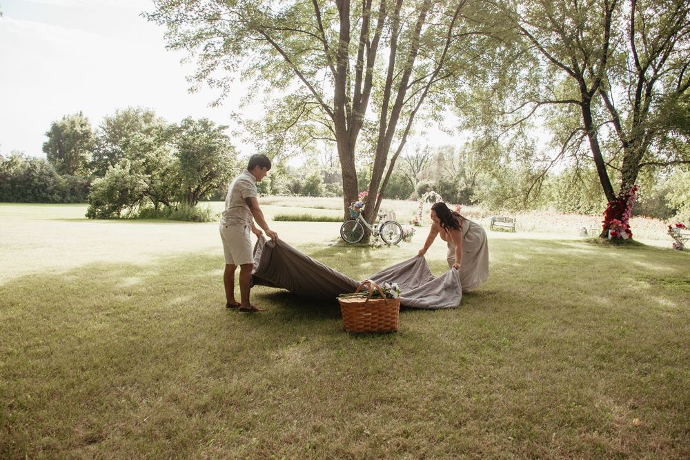 Two people set up a summer picnic blanket with a basket on a sunny grassy lawn beneath tall trees.