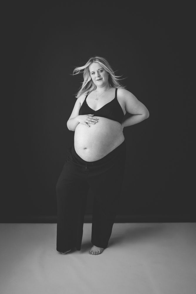 A black and white maternity photograph showing a plus size pregnant mother against a black background and white floor wearing a black bra.