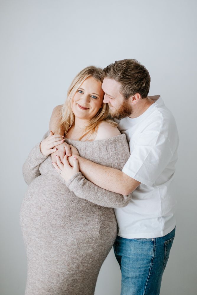 Expectant mother in a beige jumper dress is hugged by her partner while he looks over her shoulder to her bump.