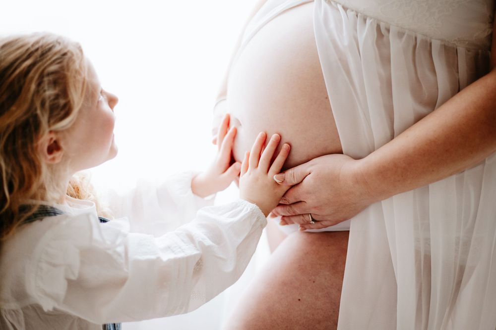 Small child touching pregnant belly wearing white dress while mum wears flowy maternity gown in soft natural light.