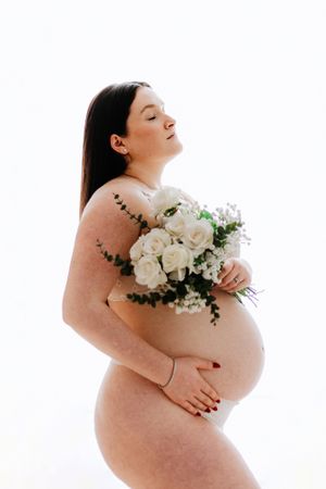 An artistic maternity photoshoot featuring white roses and baby's breath flowers against a bright background.
